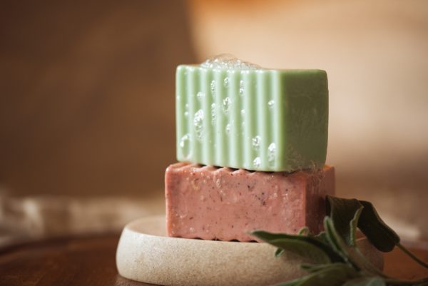Group of two chakra soaps - Root Chakra Soap and Heart Chakra Soap, with bubbles, sitting on a stone soap dish with herbs | Shine Body & Bath